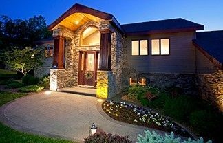 A stone-accented home exterior at twilight, featuring a lighted entryway, stone columns, and a curved paved patio.