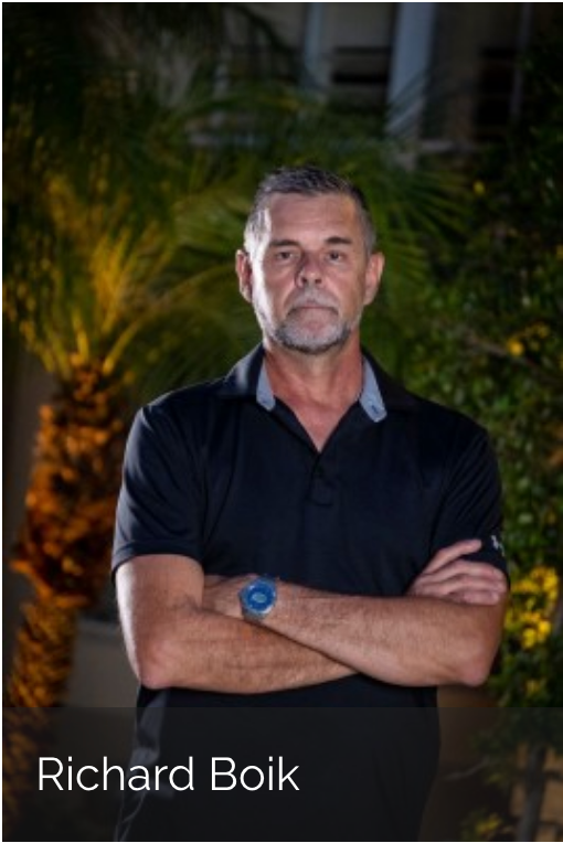 Richard Boik stands outdoors at night with arms crossed, wearing a black polo shirt, with foliage in the background.