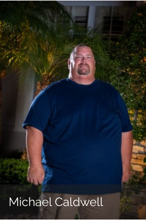 Michael Caldwell stands outdoors at night wearing a dark blue t-shirt and khaki pants against a backdrop of foliage.