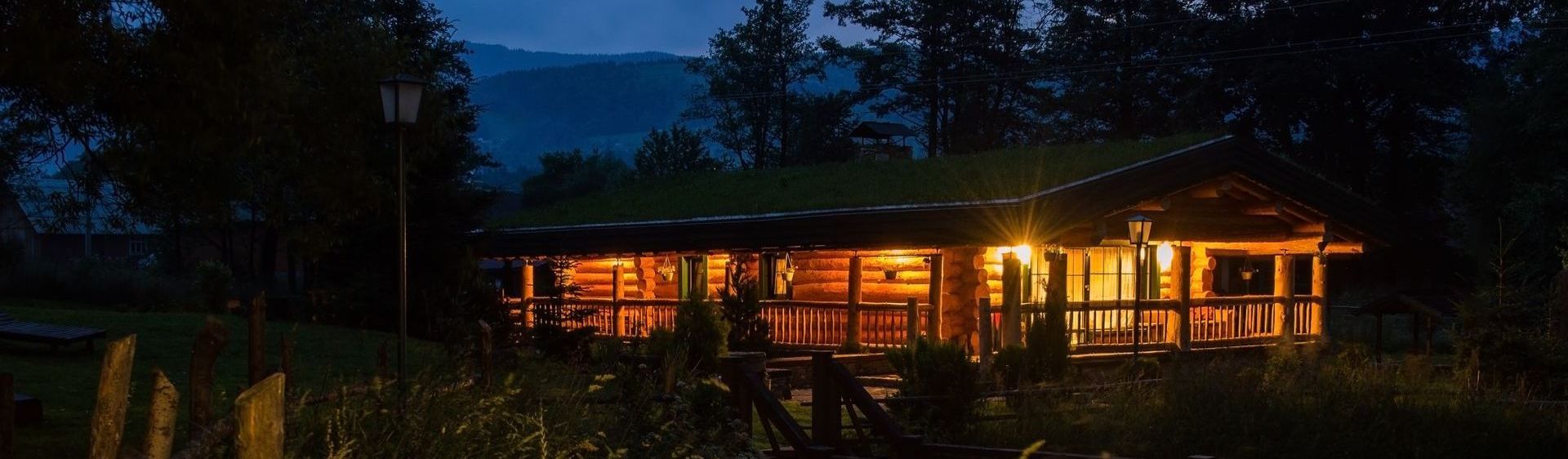 A cozy, illuminated log cabin at twilight, nestled in a landscape with silhouettes of trees and distant mountains.