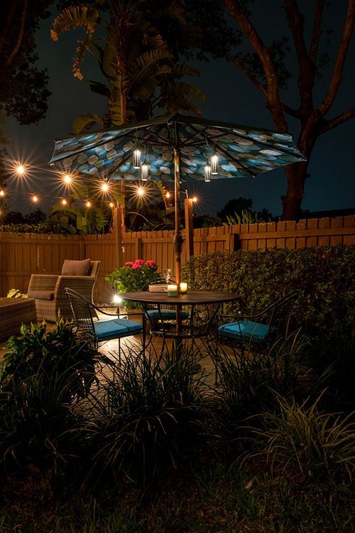 A backyard patio at night features a table, chairs, an illuminated umbrella, and string lights against a wooden fence.