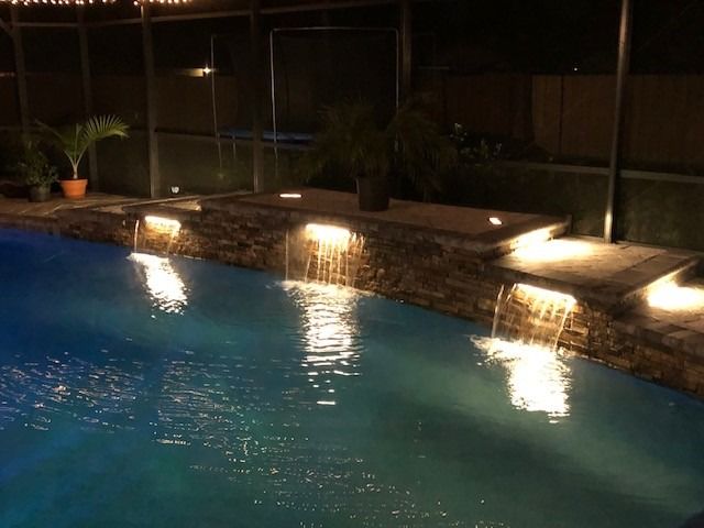 A glowing swimming pool with stone waterfall features illuminated by warm, soft lighting at night.