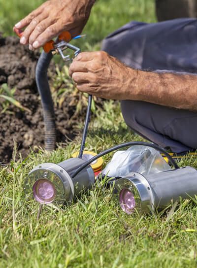 A person kneels in the grass, adjusting a wire connected to two circular, metal, pink-lensed lights on the ground.