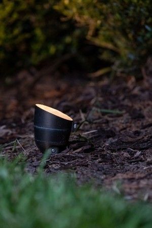 A small, black, angled landscape spotlight glows on dark mulch near a blurred green bush.