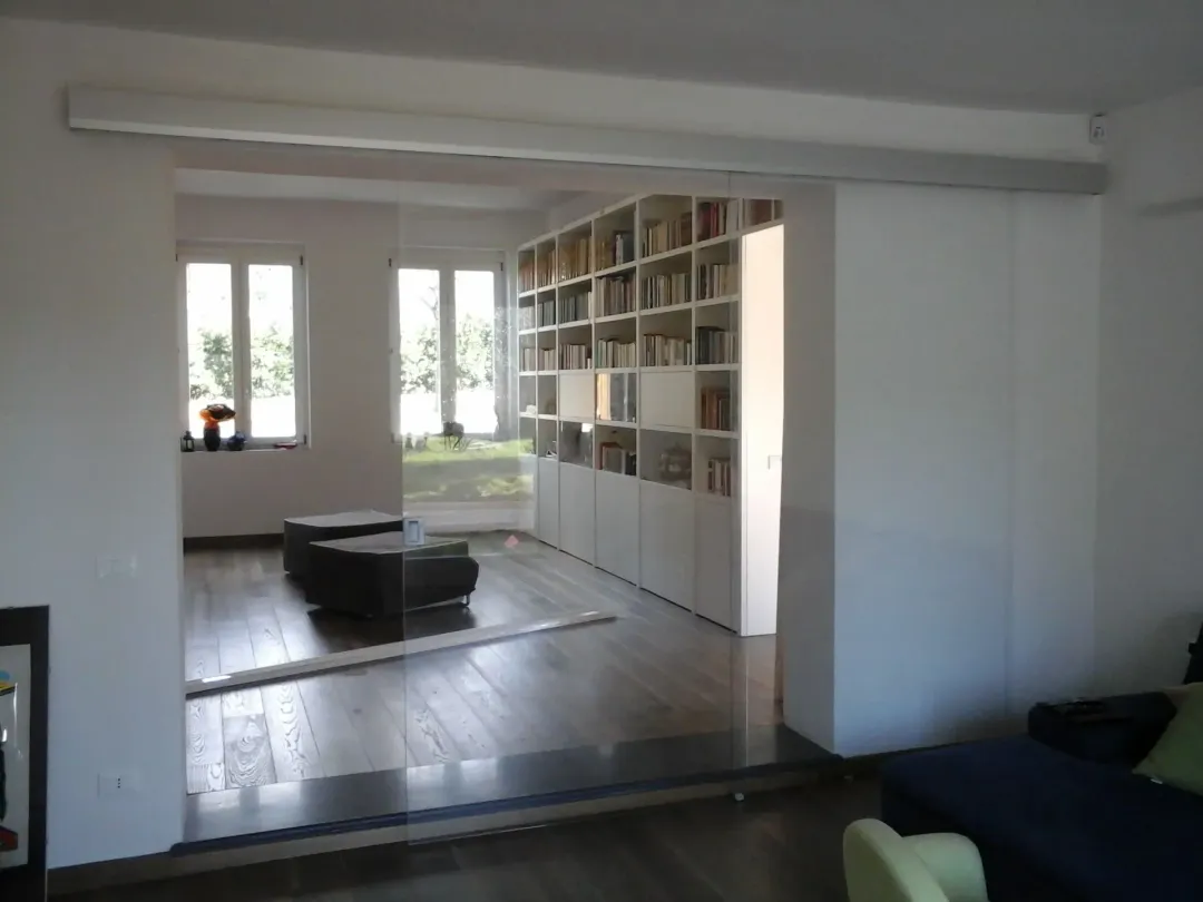 Soggiorno moderno con pareti bianche, pavimento in legno, scaffali a muro e un divano nero vicino a una finestra.