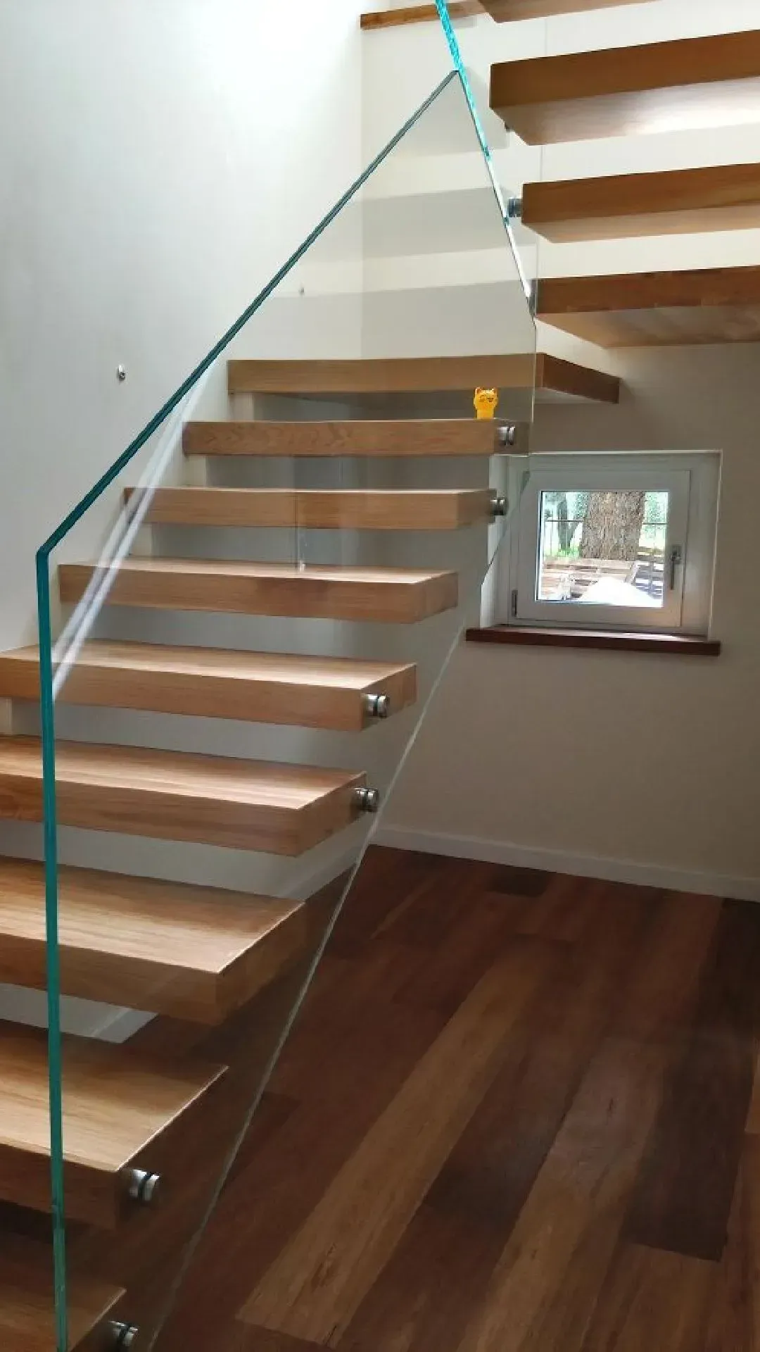 Scala moderna con gradini in legno, ringhiera in vetro e una piccola finestra sopra un pavimento in legno massello.