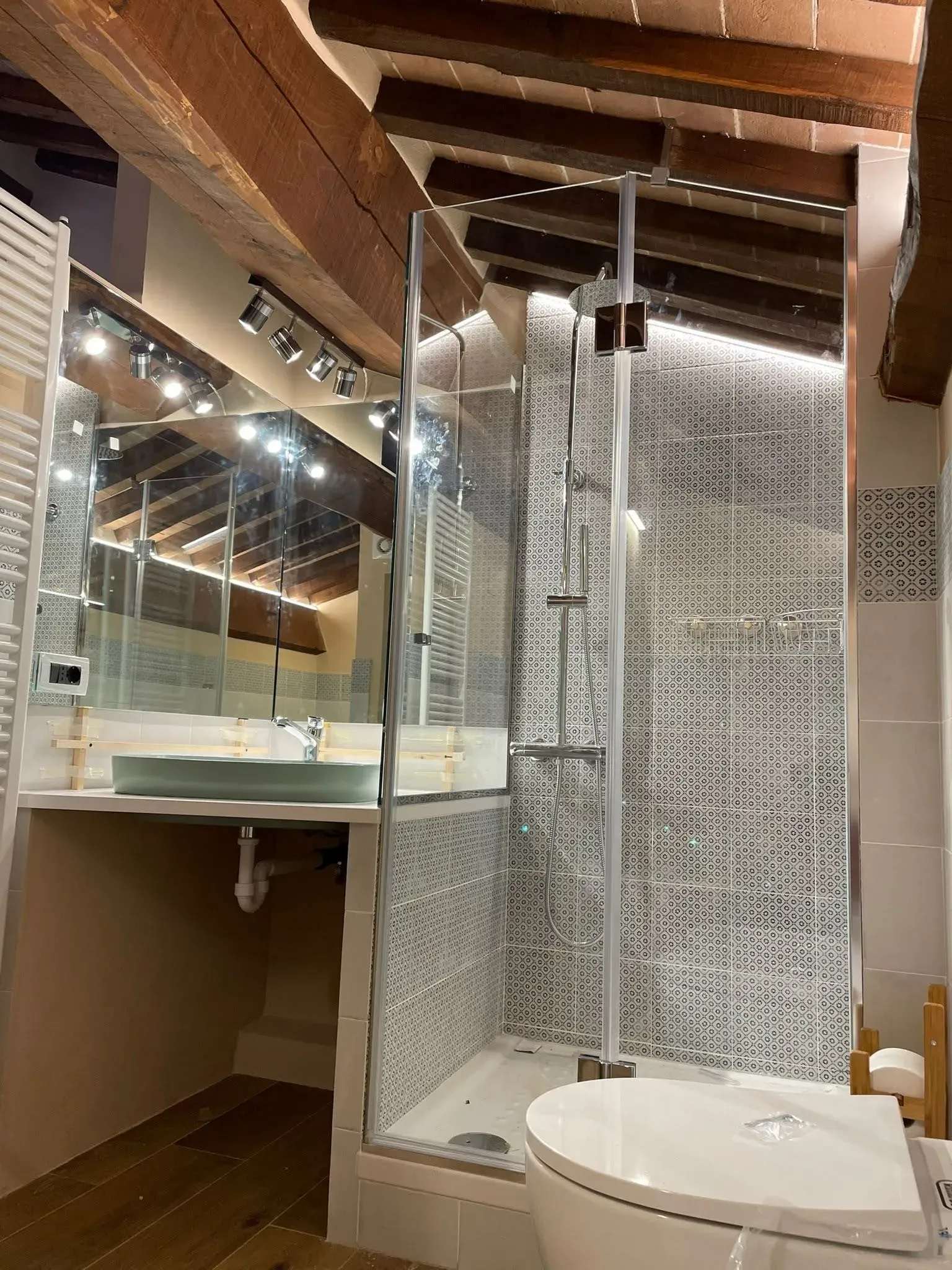 Bagno moderno con doccia in vetro, lavabo bianco, piastrelle decorate e travi in ​​legno a vista sul soffitto.