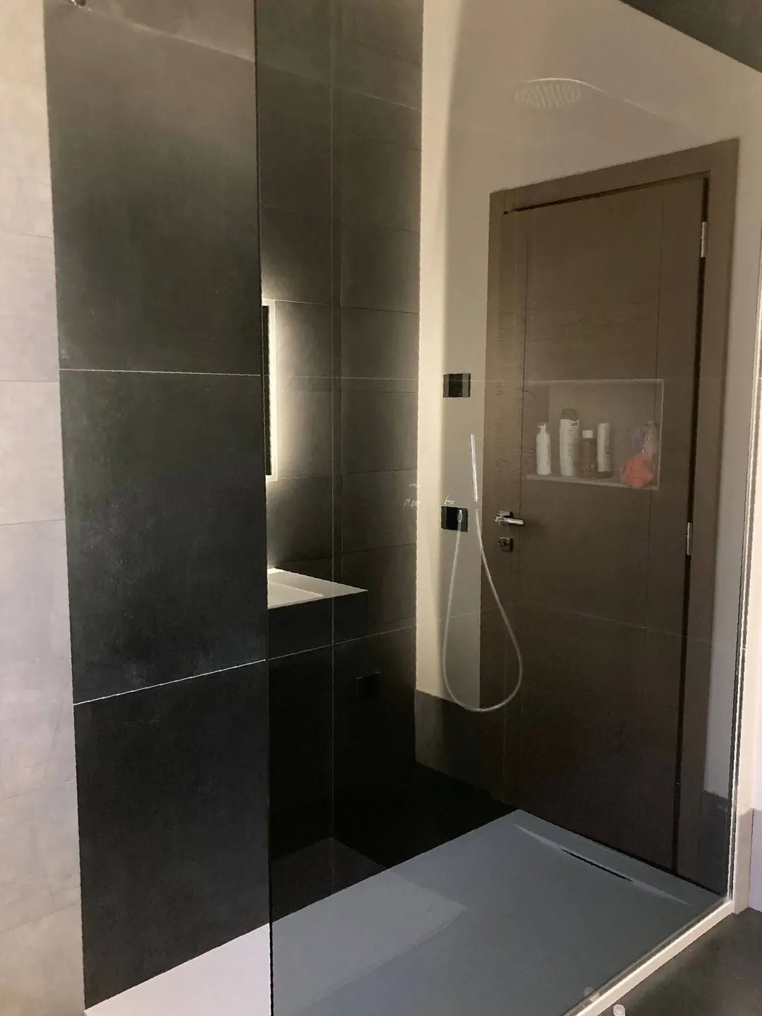 Bagno moderno con doccia piastrellata di scuro, mobile lavabo nero e porta grigia chiusa.