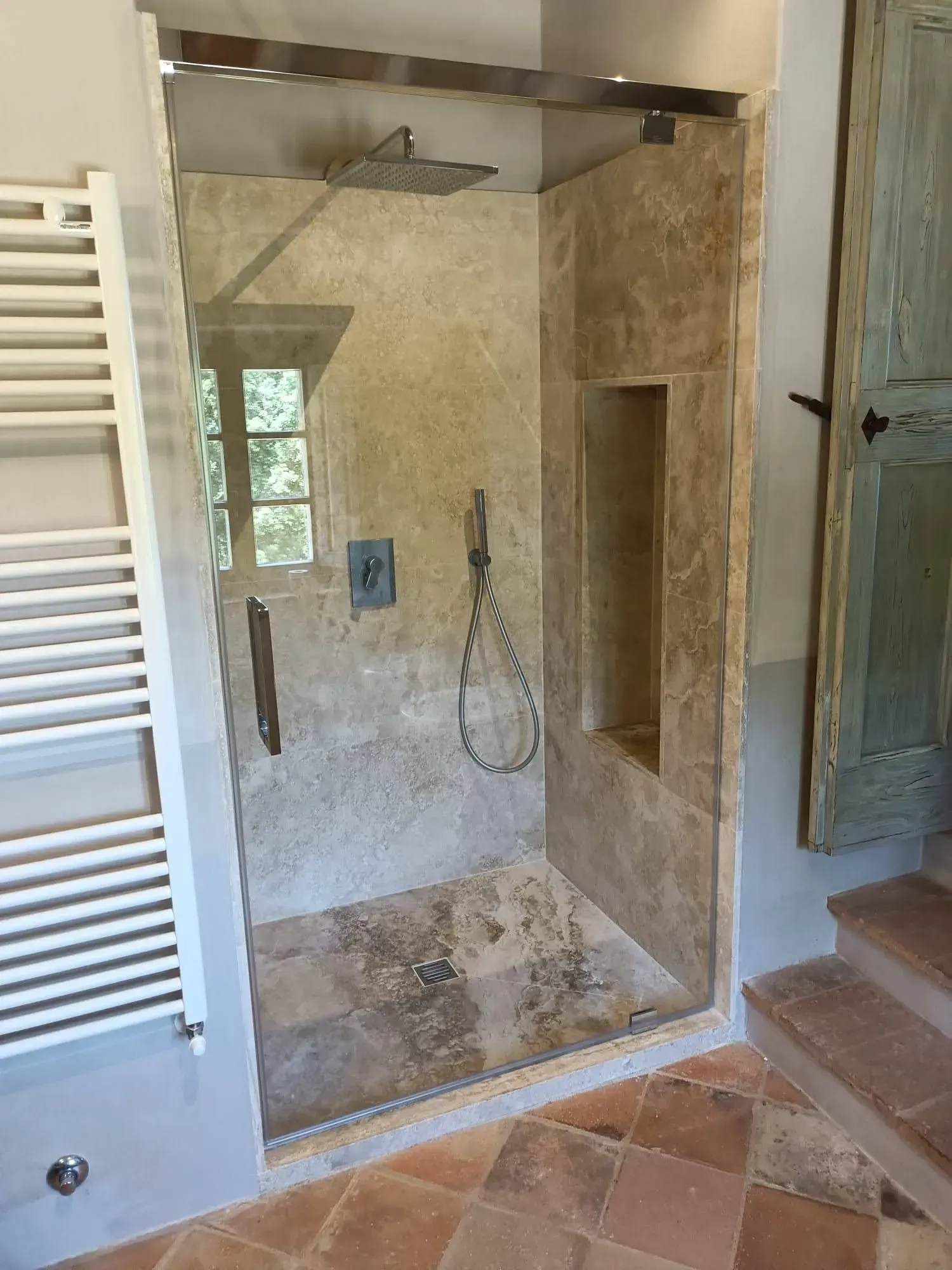 Cabina doccia con piastrelle beige, porta in vetro e nicchia integrata accanto alle piastrelle del pavimento in terracotta.