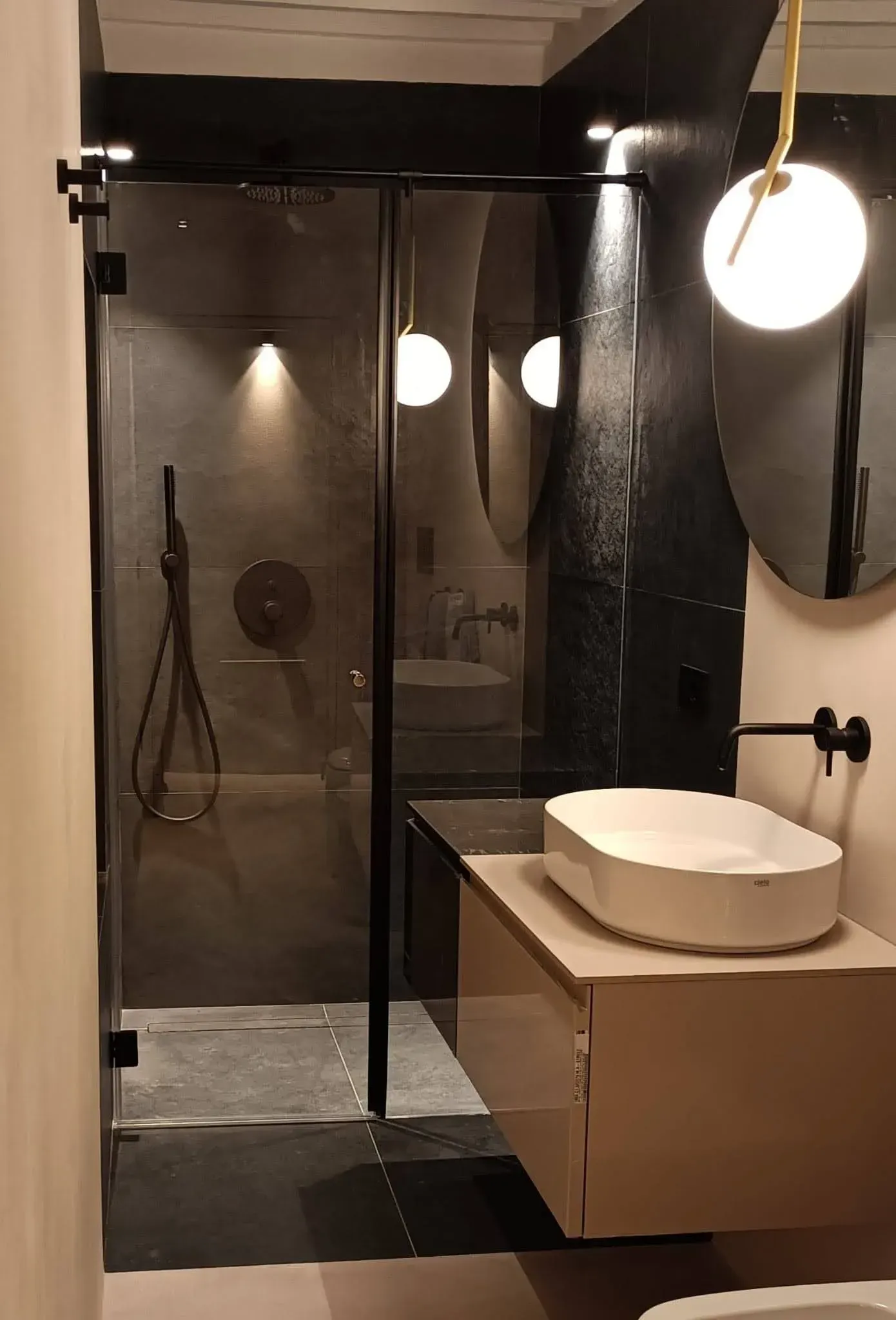 Bagno moderno con doccia in vetro, pareti nere e mobile lavabo sospeso in legno con lavabo rotondo.