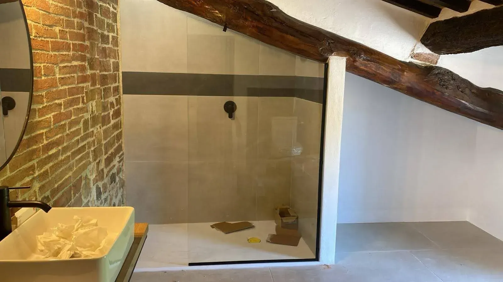 Bagno mansardato in fase di ristrutturazione con parete in mattoni, zona vasca/doccia bianca e travi in ​​legno a vista.