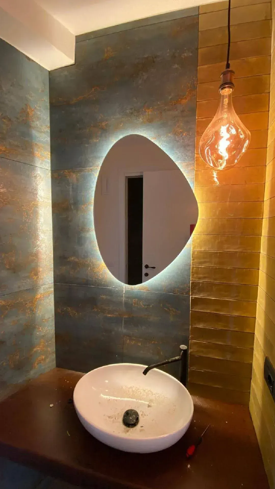 Bagno moderno con specchio ovale, lavabo bianco, parete in legno caldo e lampada a sospensione color ambra.