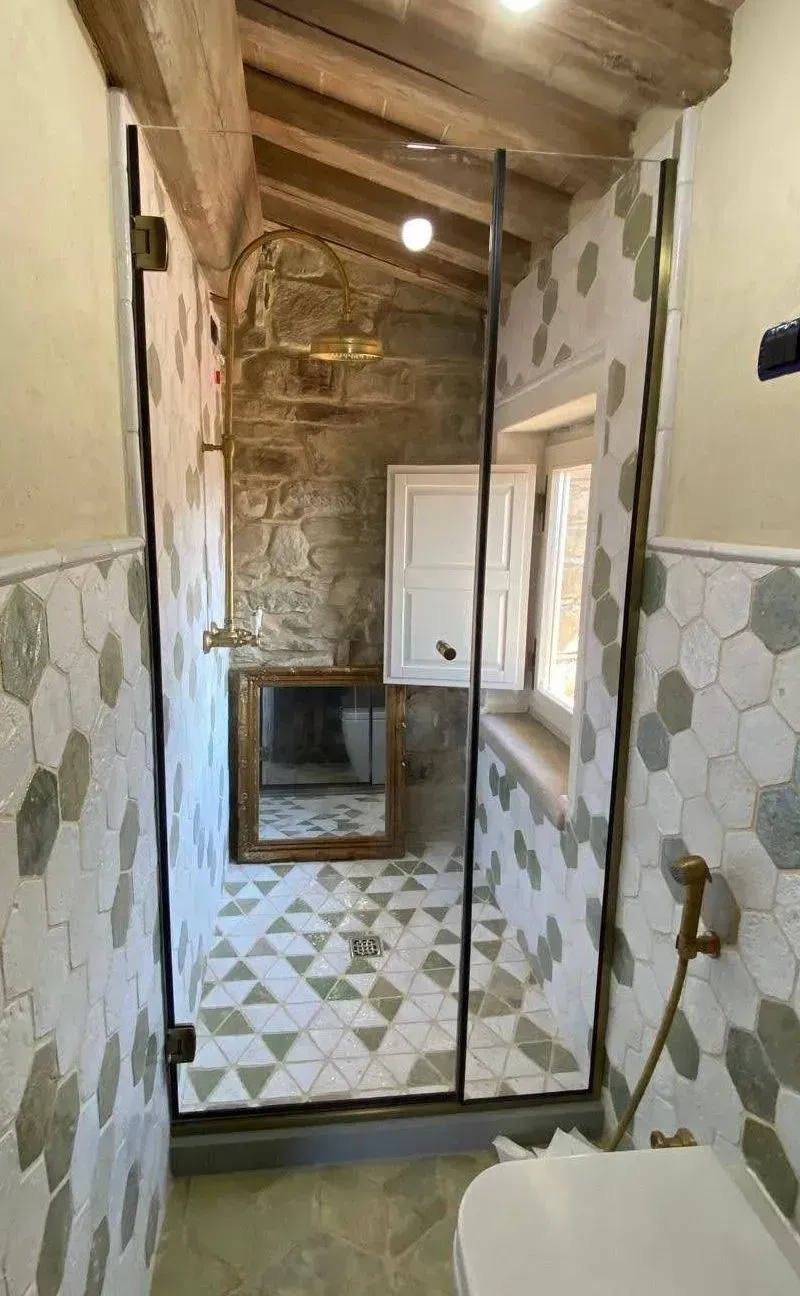 Stretto corridoio del bagno piastrellato con pareti in pietra, soffitto in legno, specchio e piccolo lavandino in fondo.