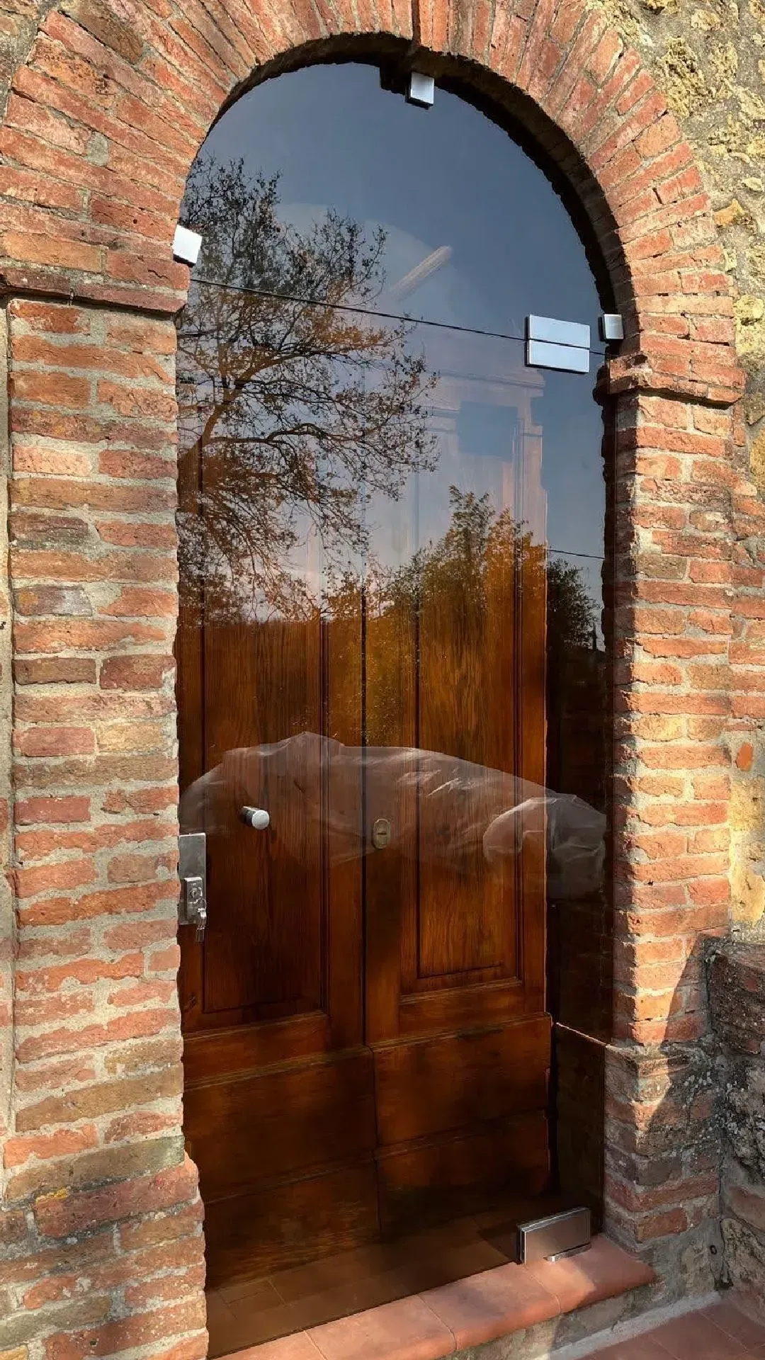 Porta ad arco in legno e vetro incastonata in un muro di mattoni, che riflette alberi e cielo.