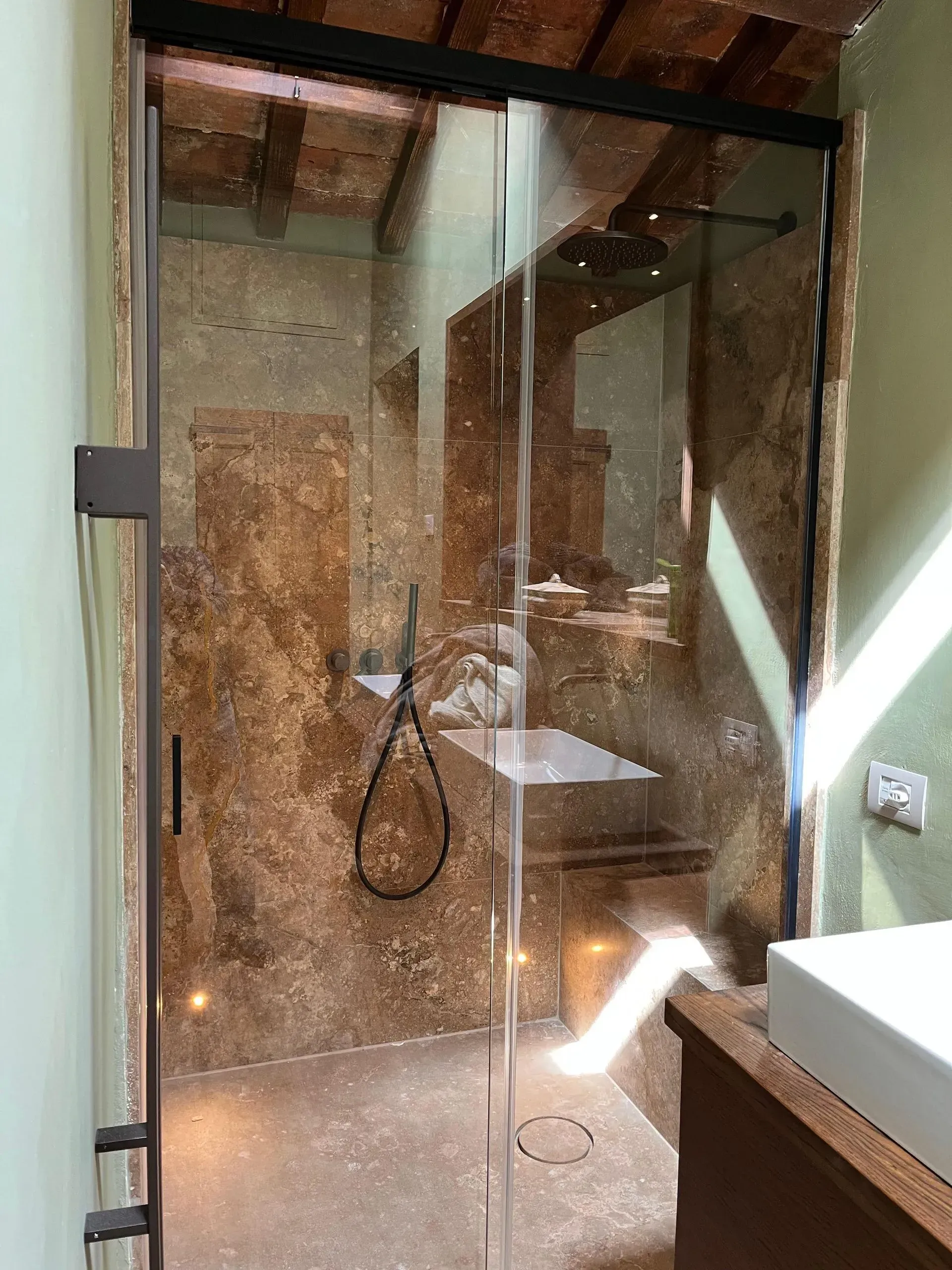 Box doccia in vetro con pareti in pietra, soffione a pioggia e panca in un bagno piastrellato.
