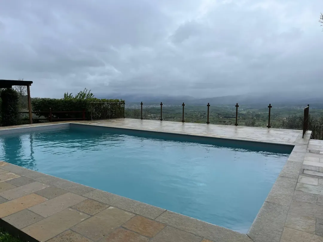 Piscina all'aperto con acqua turchese che si affaccia su un paesaggio nuvoloso e sulle colline in lontananza.
