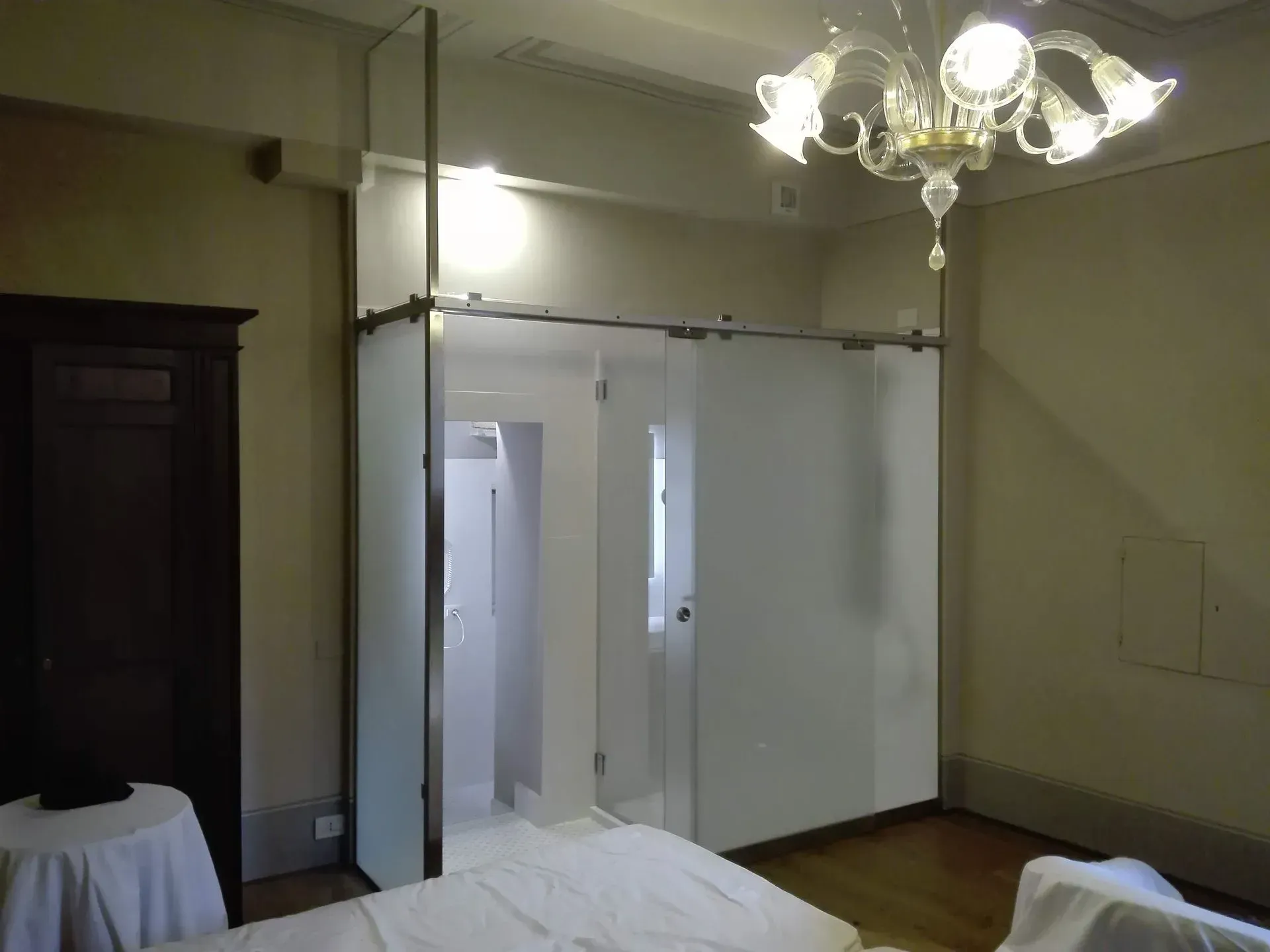 Stanza vuota con un lampadario, porte scorrevoli bianche divisorie, un armadio scuro e un tavolo ricoperto da una tovaglia bianca.