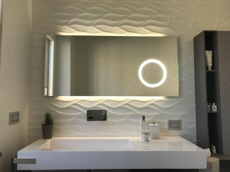 Mobile bagno moderno con specchio a parete, lampada circolare, lavabo bianco e rivestimento a parete con piastrelle texturizzate.