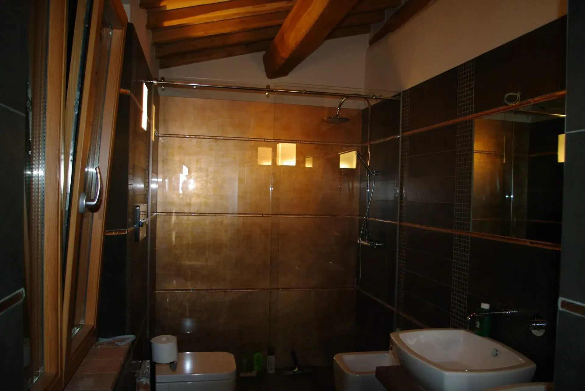 Bagno moderno con doccia in vetro, pareti rivestite con piastrelle scure e travi a vista sul soffitto.