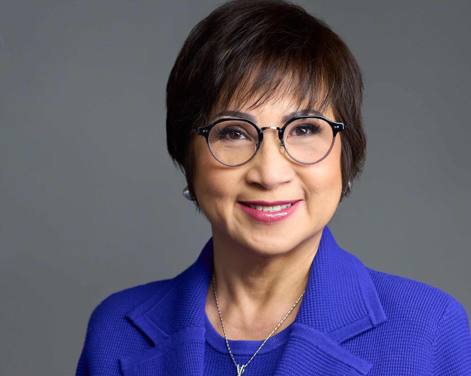 A woman wearing glasses and a blue jacket is smiling for the camera.