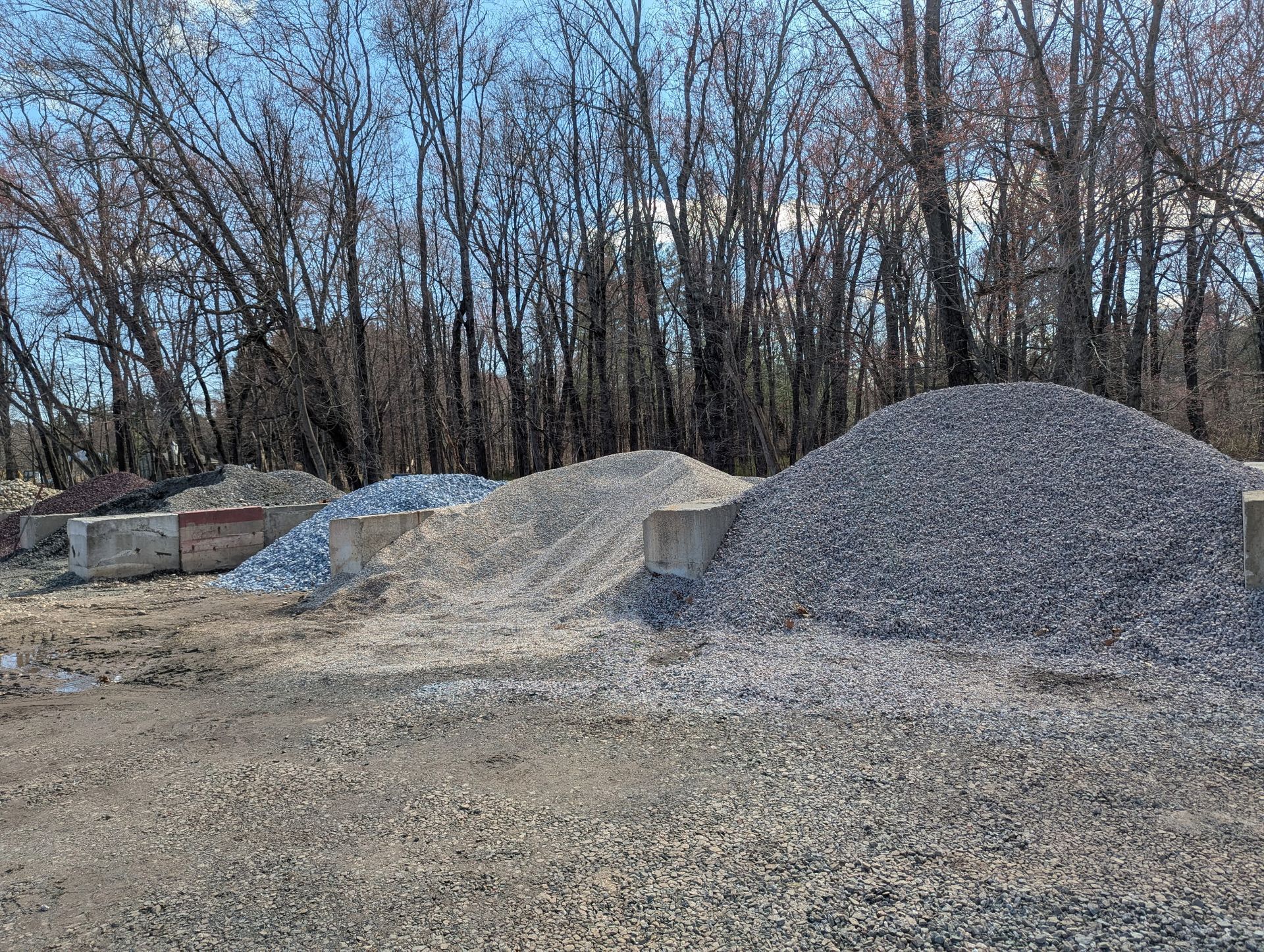 Piles of gray crushed gravel and stone sit behind concrete barriers in a yard with a background of bare trees.