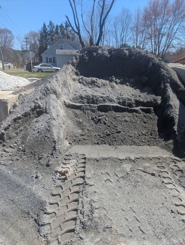 A large mound of dark gray dirt and gravel at a construction site, with deep tire tracks visible in the foreground.