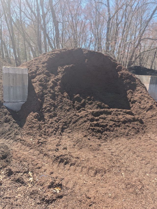 A large pile of dark brown wood mulch sits between two concrete block retaining walls outdoors in a wooded area.