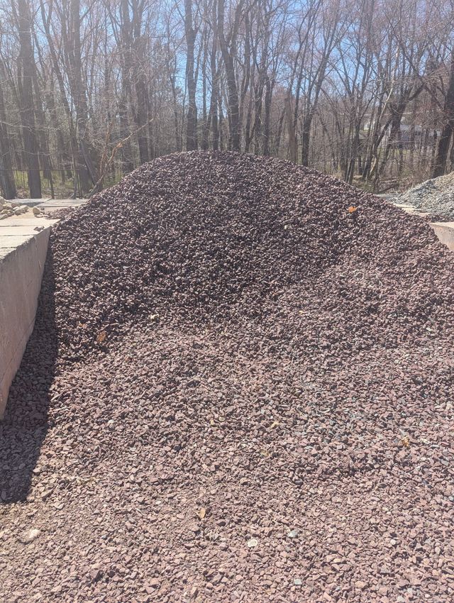 A large, conical pile of dark brown crushed landscaping stone outdoors against a background of bare trees.