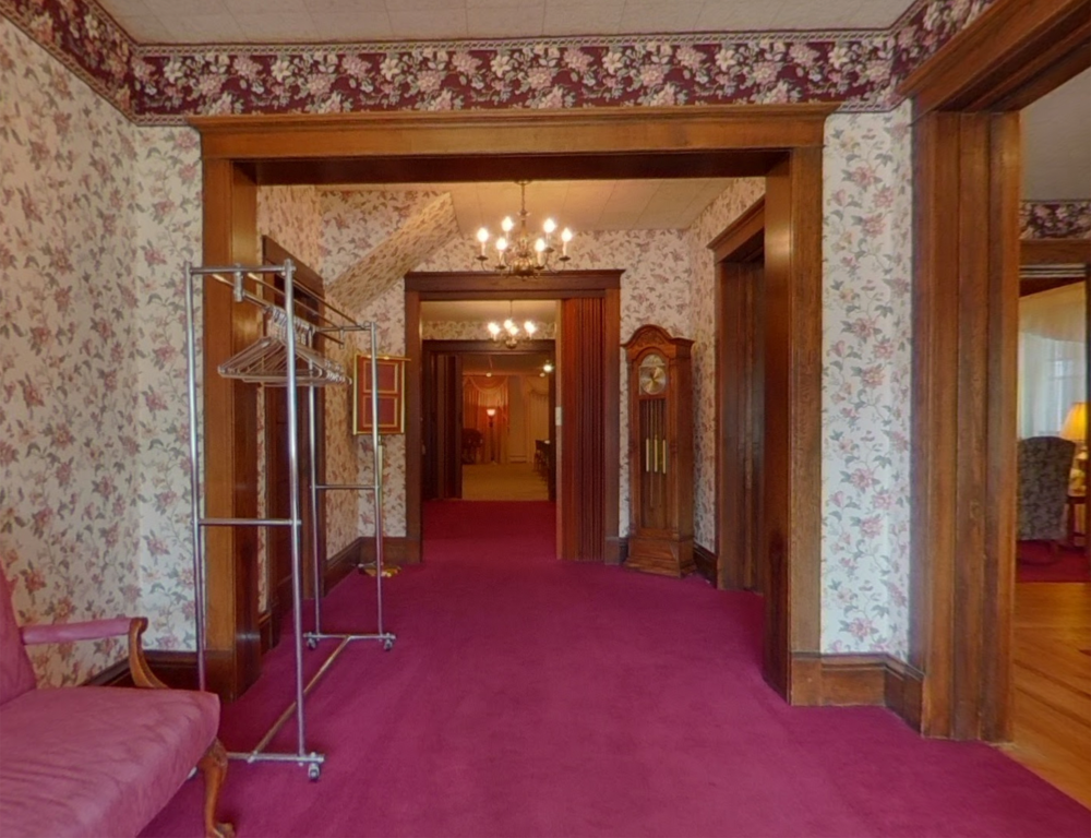 A hallway with floral wallpaper, a magenta carpet, a rolling garment rack, and a wooden grandfather clock.