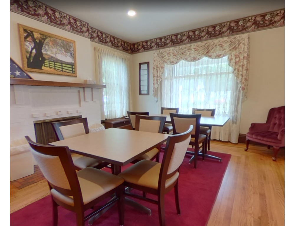 A dining room with two square tables, wood chairs with beige cushions, a red rug, hardwood floors, and a corner armchair.