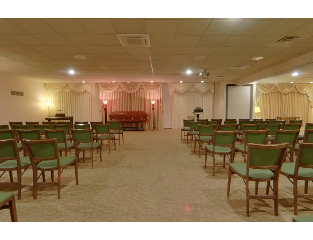 A funeral chapel interior with rows of green chairs facing a central, draped casket under soft lighting.