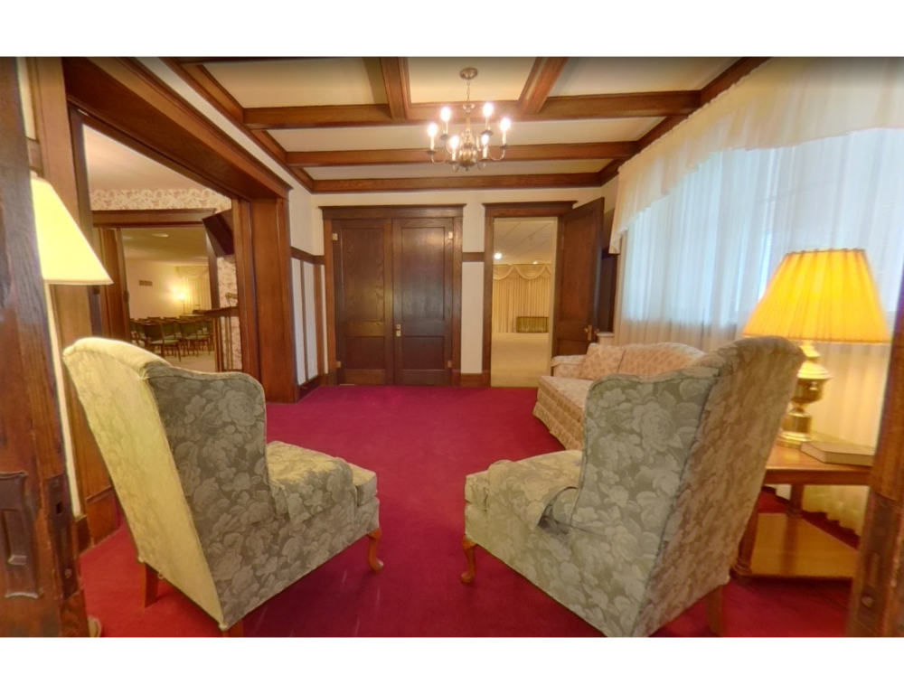 A formal living room with two patterned armchairs, a sofa, red carpeting, wood-paneled walls, and a beamed ceiling.