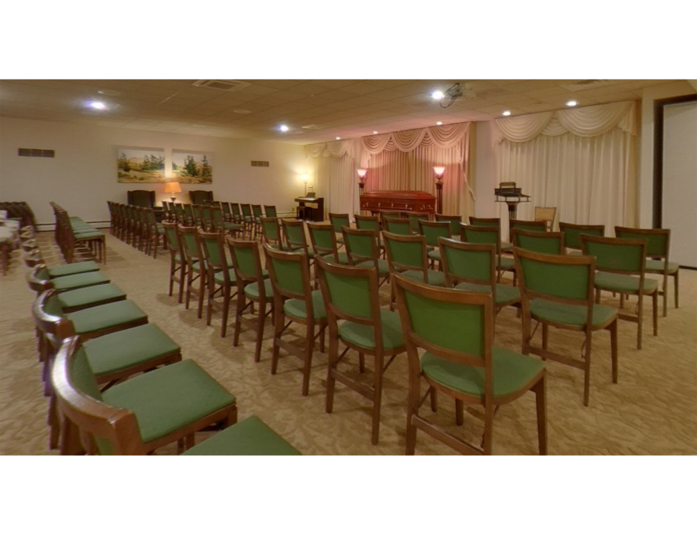 Rows of green-cushioned wooden chairs in a formal room with draped curtains, lamps, and a small stage area.