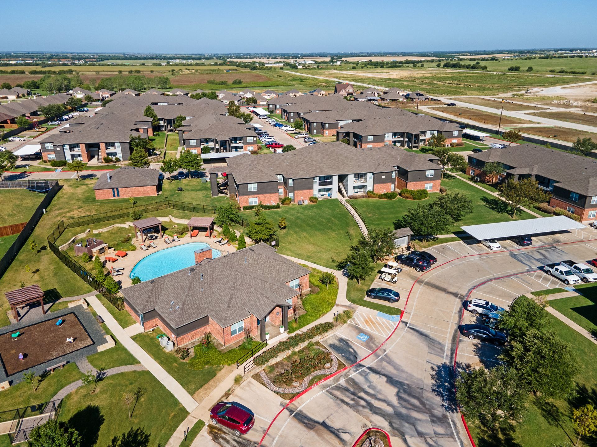 Aerial view of an apartment complex with pool and surrounding green spaces under a clear blue sky.