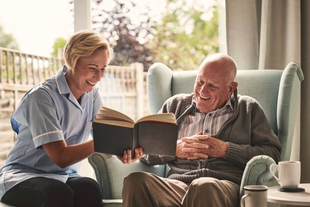 Caregiver and older adult share a book, smiling in a sunny room.
