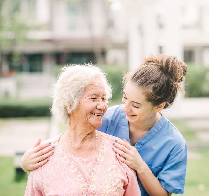 Woman smiles at a senior woman, hands on her shoulders. Outdoors, blurred background.