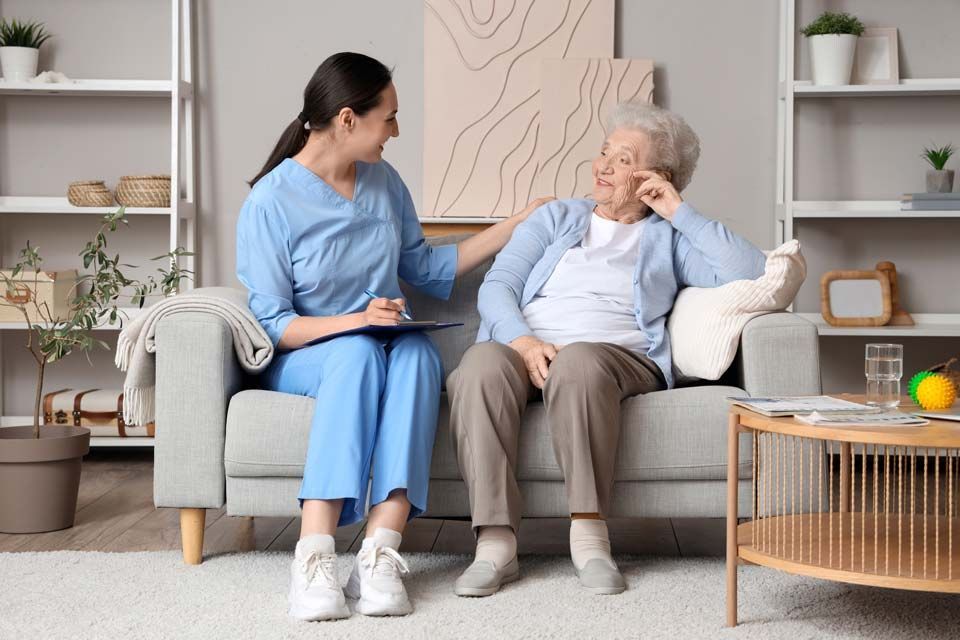 Caregiver sits beside an elderly woman on a sofa in a bright living room, speaking with her gently.