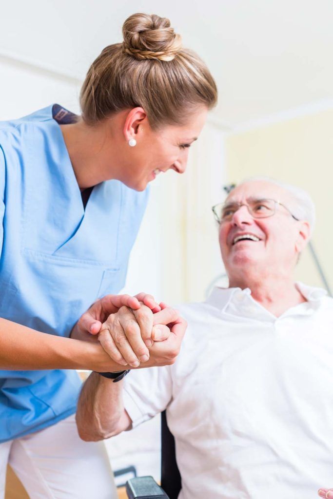 Nurse in blue scrubs assists an elderly man in a white shirt, smiling and holding his hand.