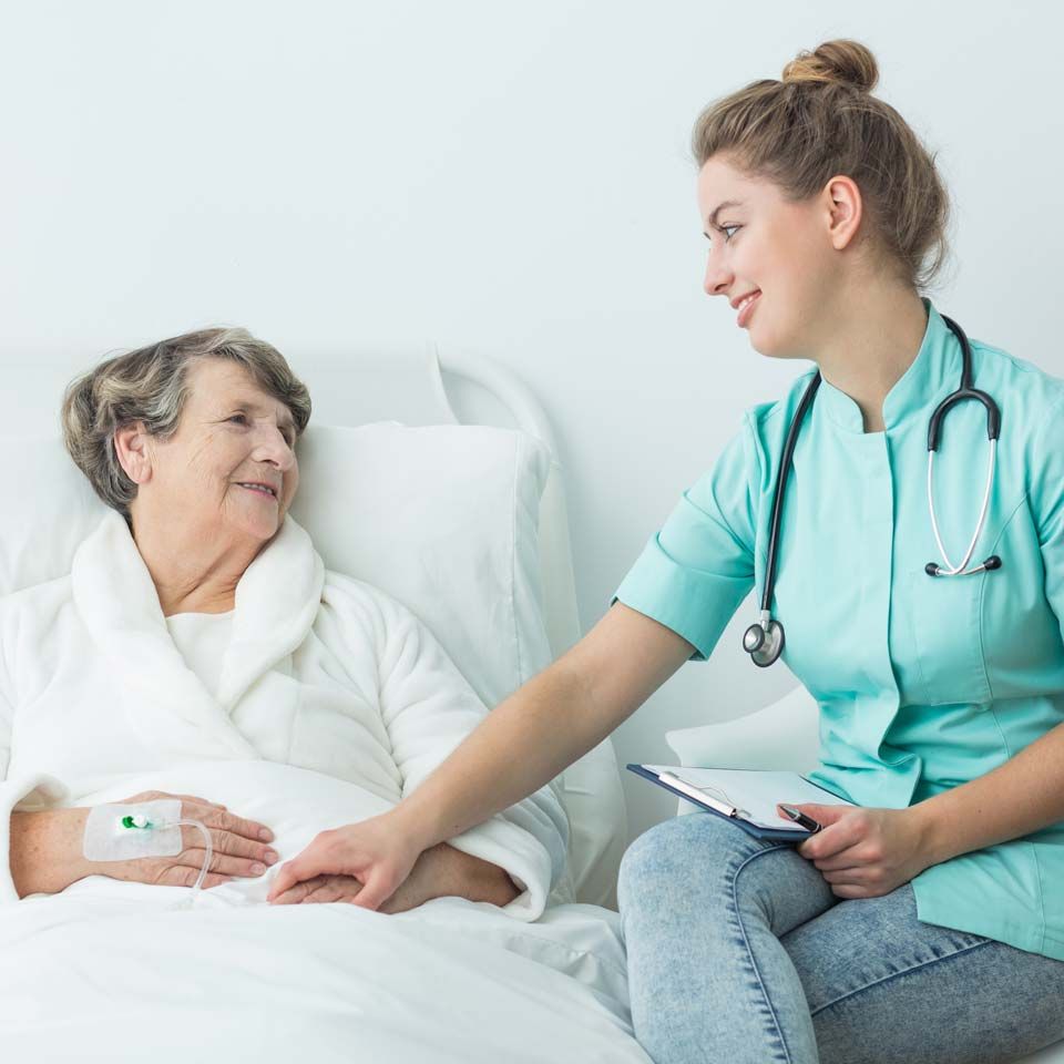 Nurse with stethoscope smiles at patient in hospital bed, hand on patient's hand.