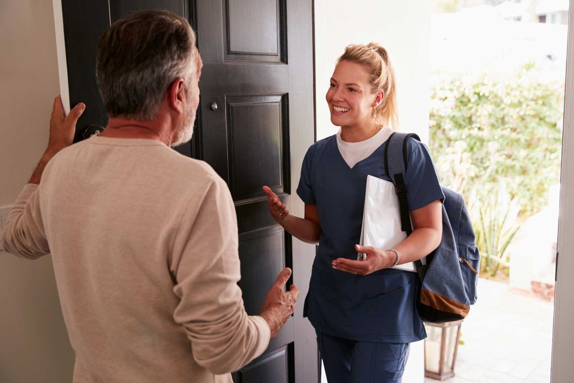 Two people talking at an open front door, one holding papers and wearing a backpack
