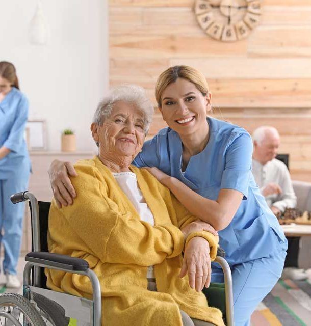 Woman in wheelchair smiles with caregiver in a well-lit room; other people in the background.