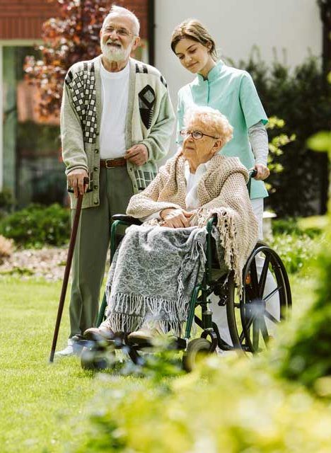 Elderly person in a wheelchair with caregiver and man in a garden.