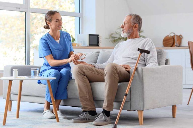 Caregiver in blue scrubs assists a person sitting on a sofa with a cane.