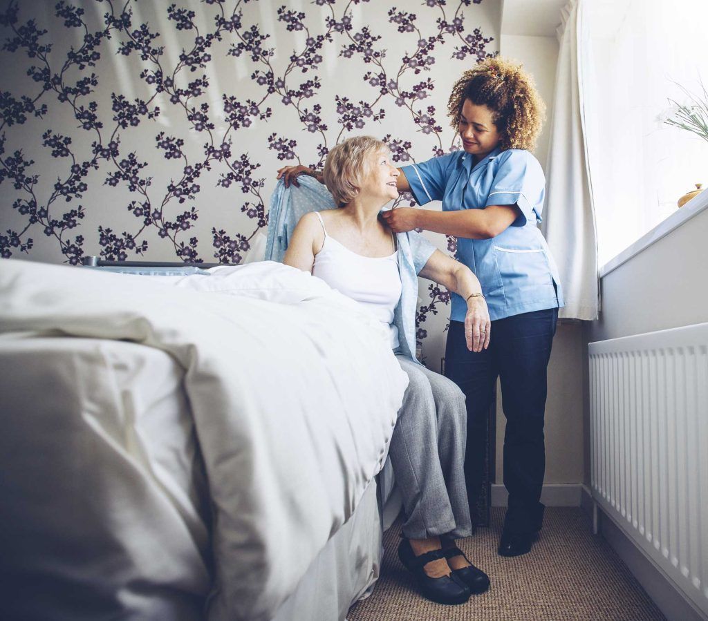 Caregiver assists a person sitting on a bed. The caregiver has their arm around them. Bright room with a window.
