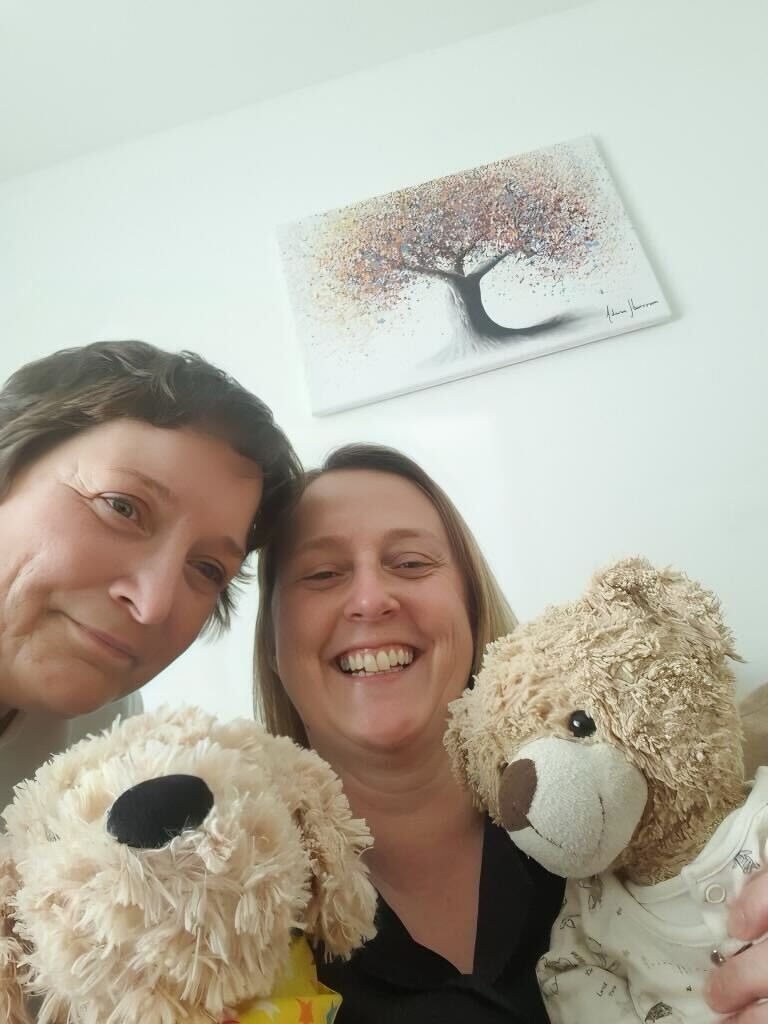Two women with stuffed animals, smiling, in front of a painting of a tree.