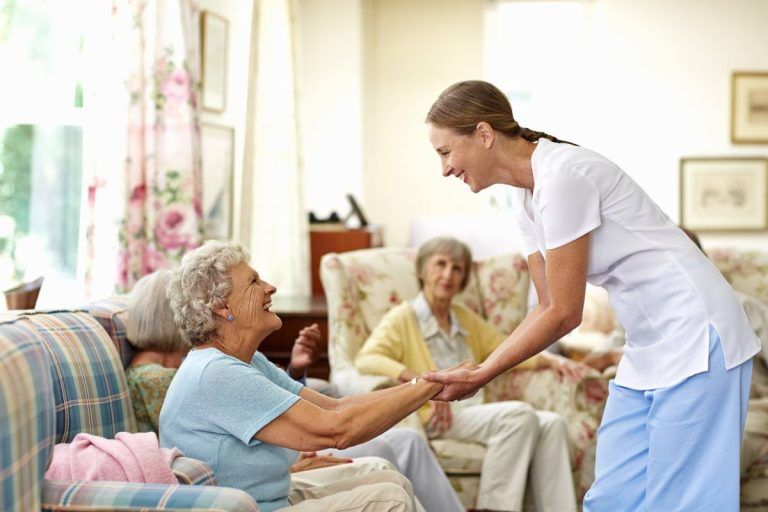 A caregiver assisting an older woman, smiling, in a brightly lit living room with other residents.