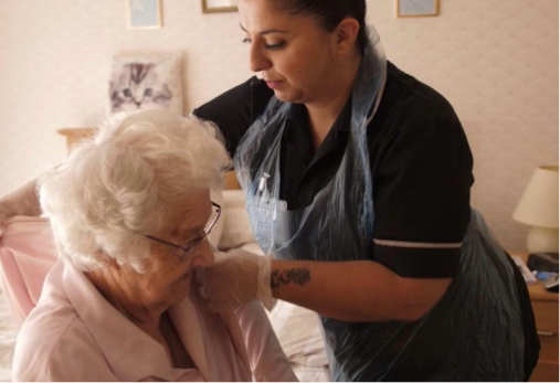 Caregiver helps an older woman with glasses in a bedroom, adjusting her clothing or arm.