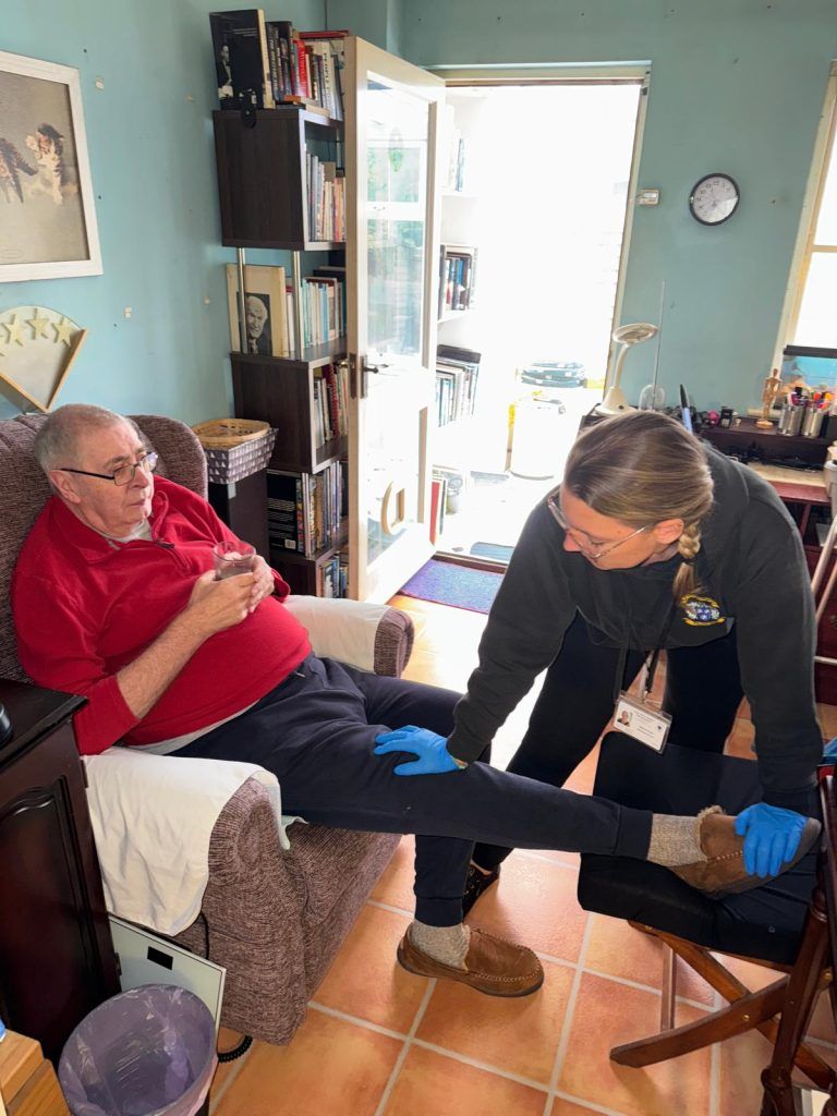 A person in a red shirt has their leg examined by a person wearing a grey shirt. Both are indoors.