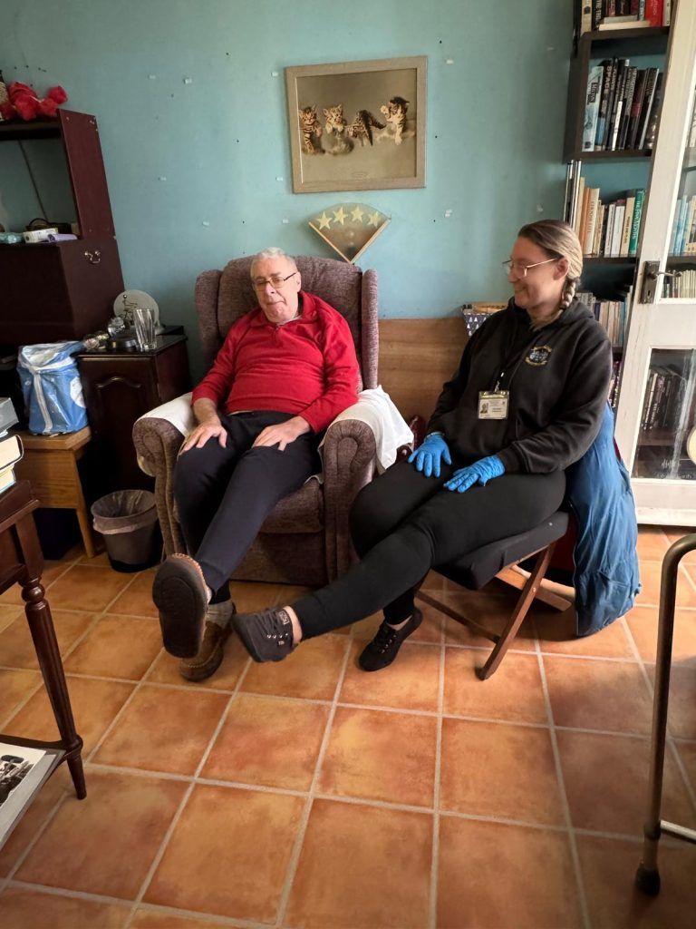 Person in red shirt sits, smiles at person wearing black jacket, gloves. Inside room, old furniture, books.