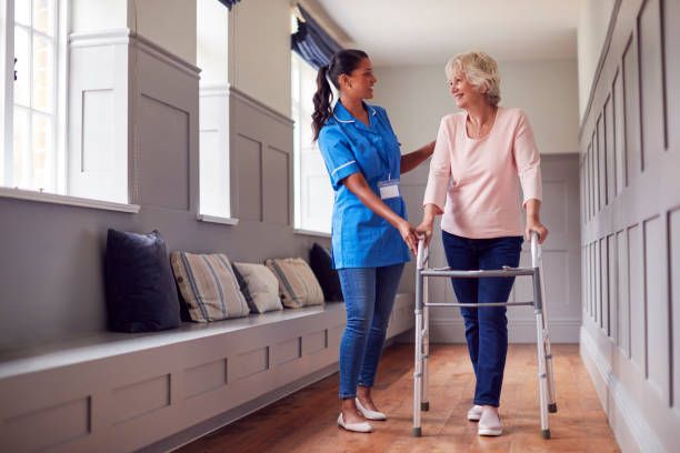 Caregiver assists a person using a walker in a hallway. The caregiver wears blue scrubs and smiles.